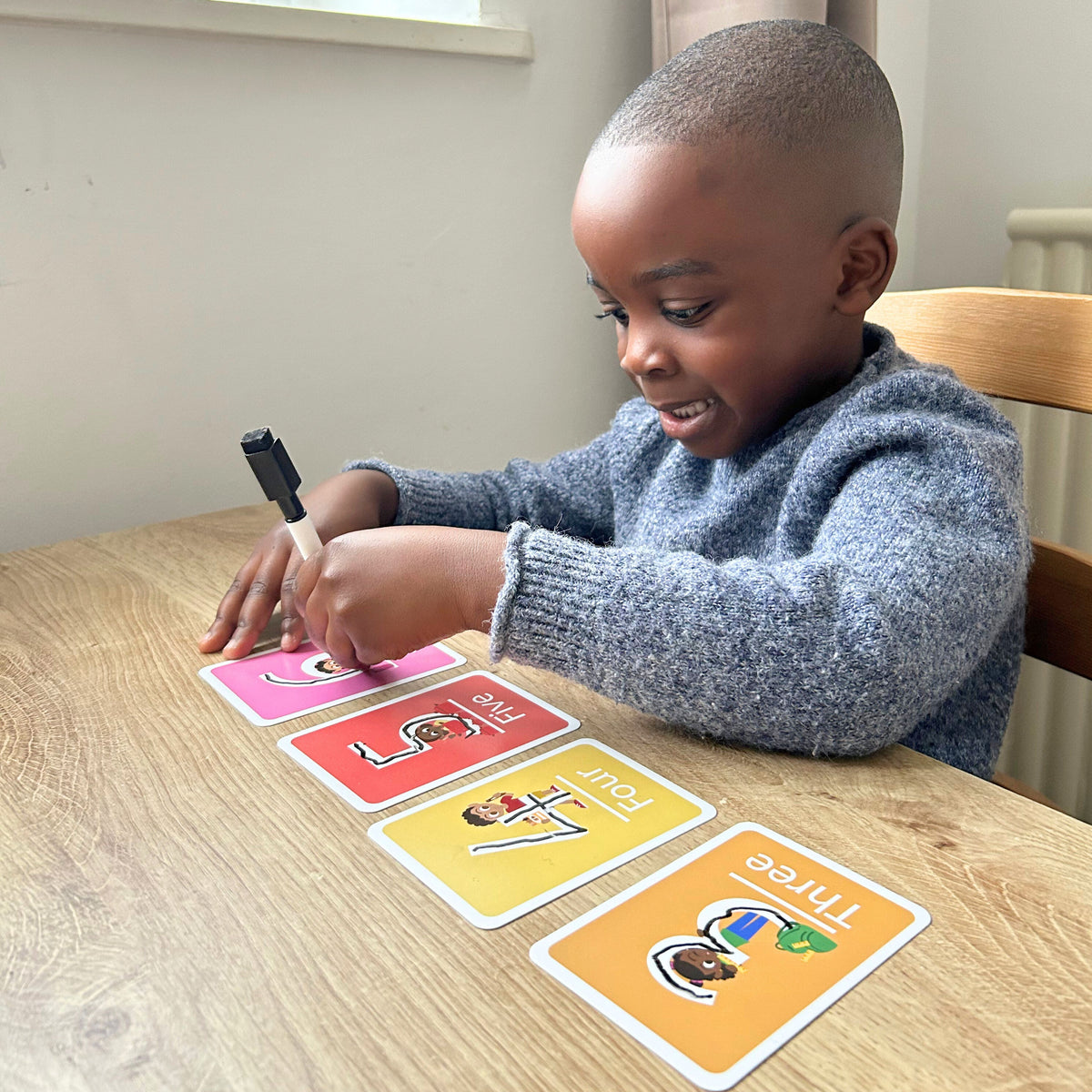 A black young boy sitting on a table with four flashcards from the Numbers set from Little Omo and he's tracing over